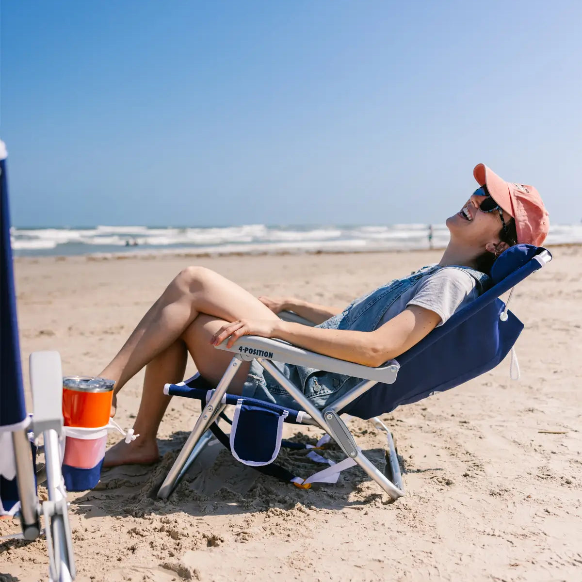 Woman reclining and laughing in a navy GCI Outdoor 4-Position Beach Chair with cupholder, enjoying the beach.