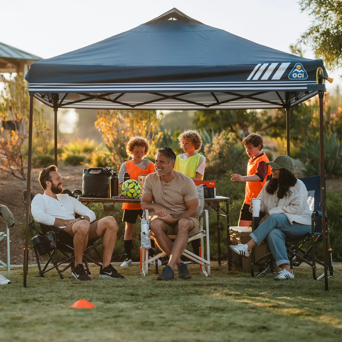 A family gathers under a dark blue LevrUp Canopy tent on a grassy lawn at sunset. Adults and children sit and stand around a small table with drinks, a cooler, and a soccer ball.