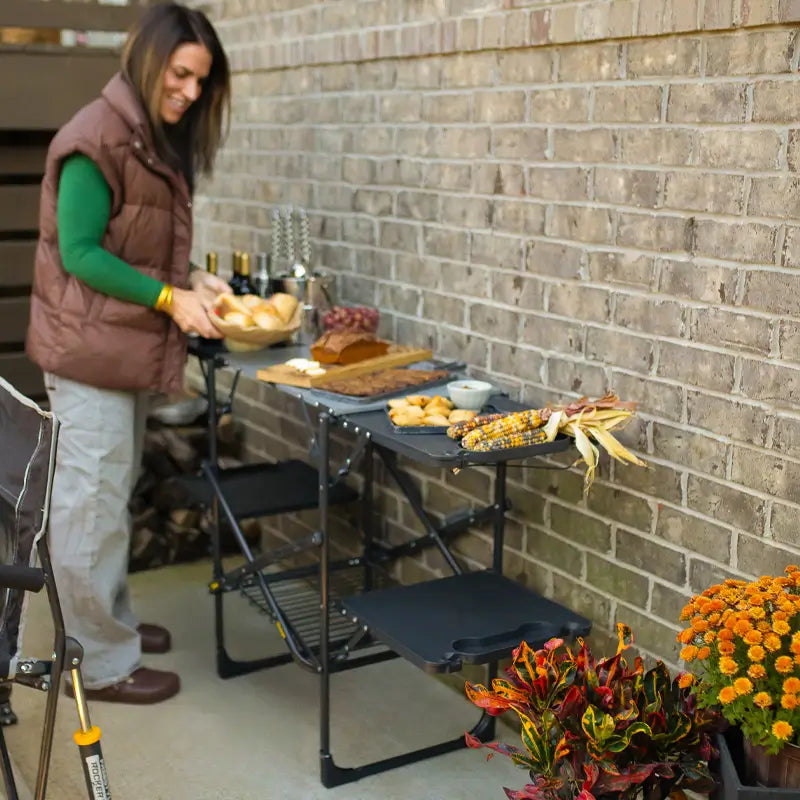 Outdoor buffet setup with food and drinks on slim-fold cook station near brick wall.