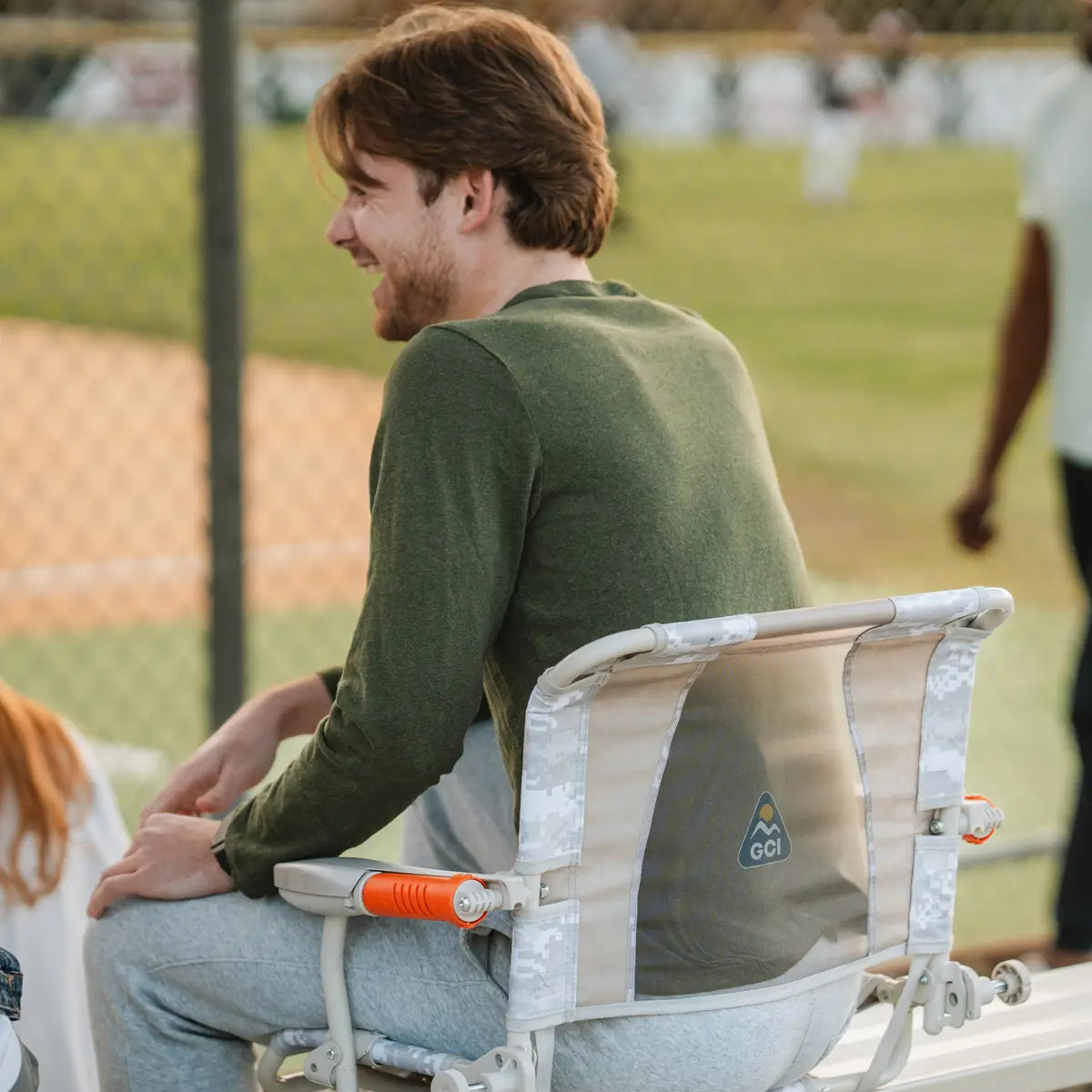 Man wearing a green shirt and gray sweatpants sits in a camo-accented GCI Outdoor stadium chair, enjoying a ballgame.