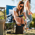 Woman sitting in a soft navy Stowaway Rocker while using a black water container with a hose attachment.