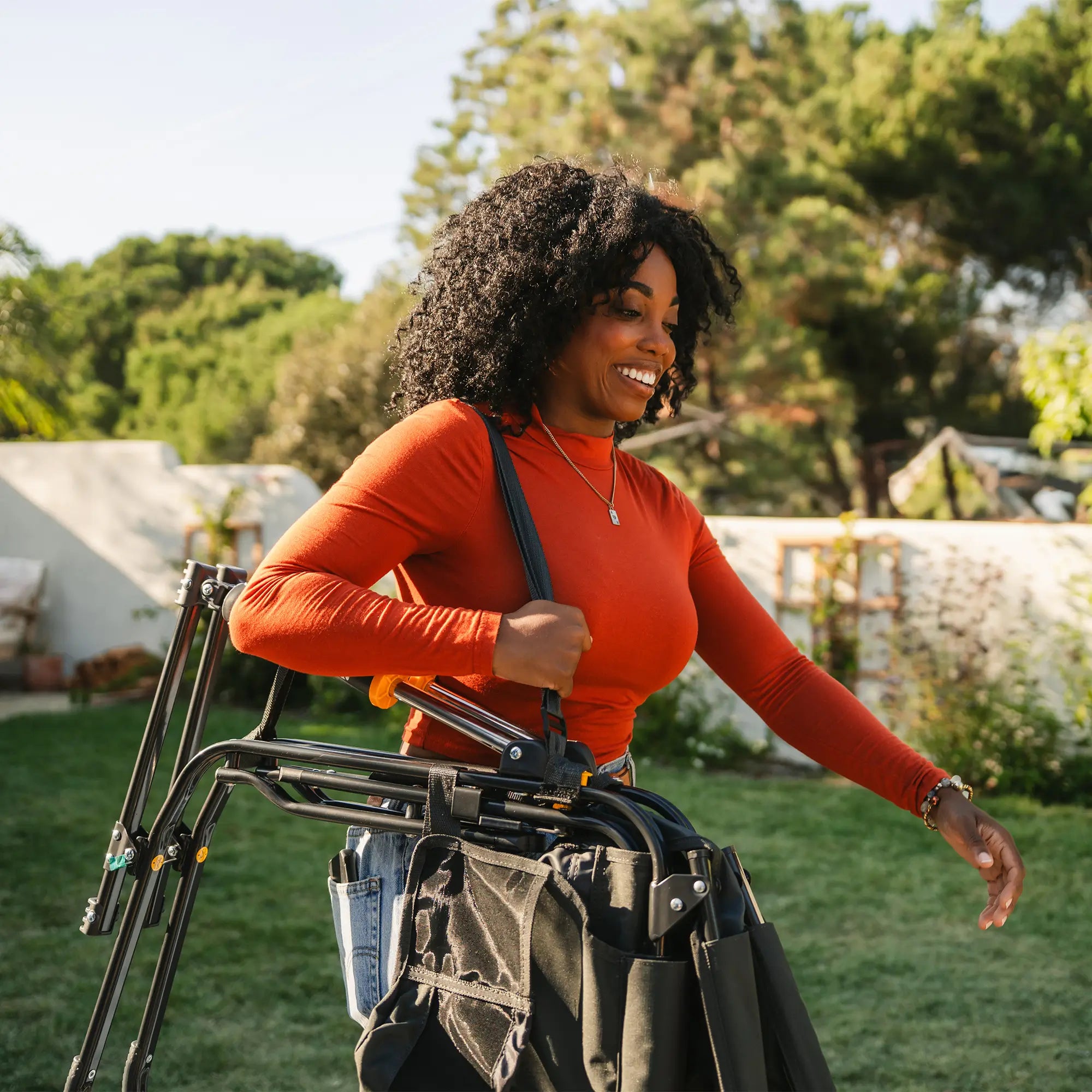 A woman walking in a backyard while carrying a black stowaway rocker around her shoulder with the attachable shoulder straps.