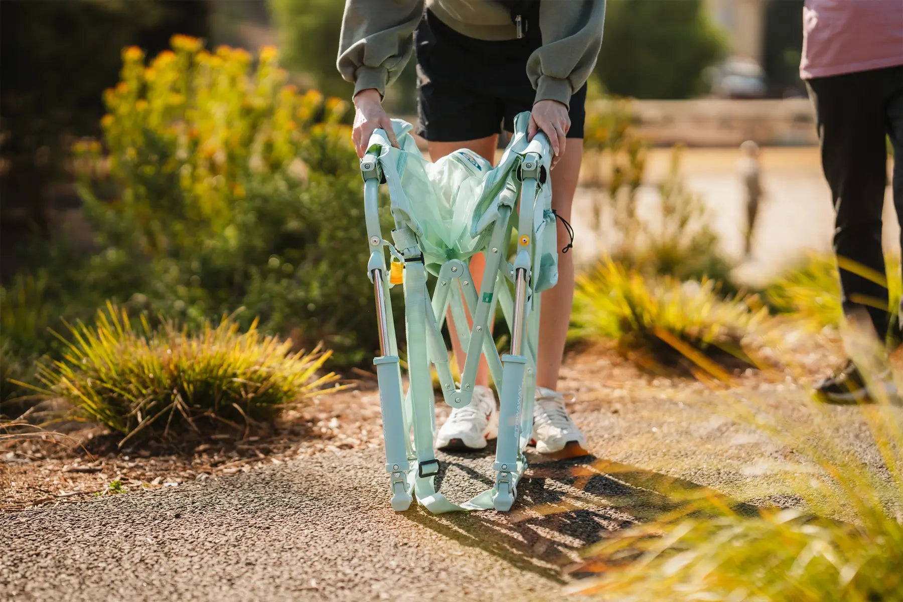 A woman collapsing a seafoam blue stowaway rocker on a gravel sidewalk.