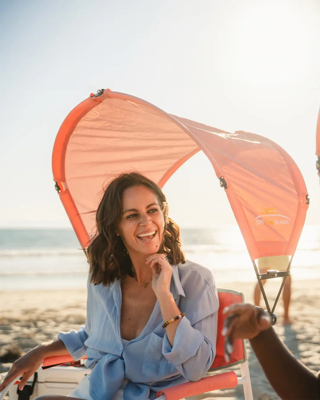 A woman sitting in a pink grab and go rocker and enjoying the beach with her attached coral sunshade accessory.