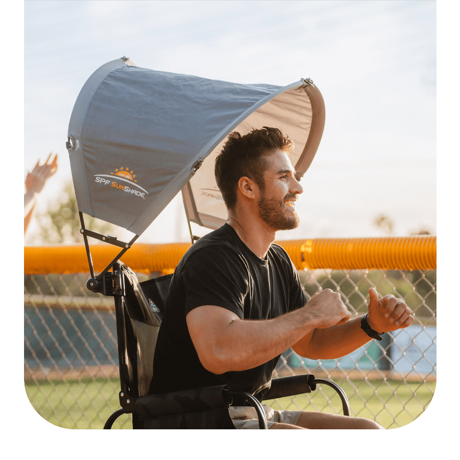 Man smiling while seated in a GCI Outdoor camp chair with an overhead SPF SunShade canopy, enjoying a sunny day near a sports field.
