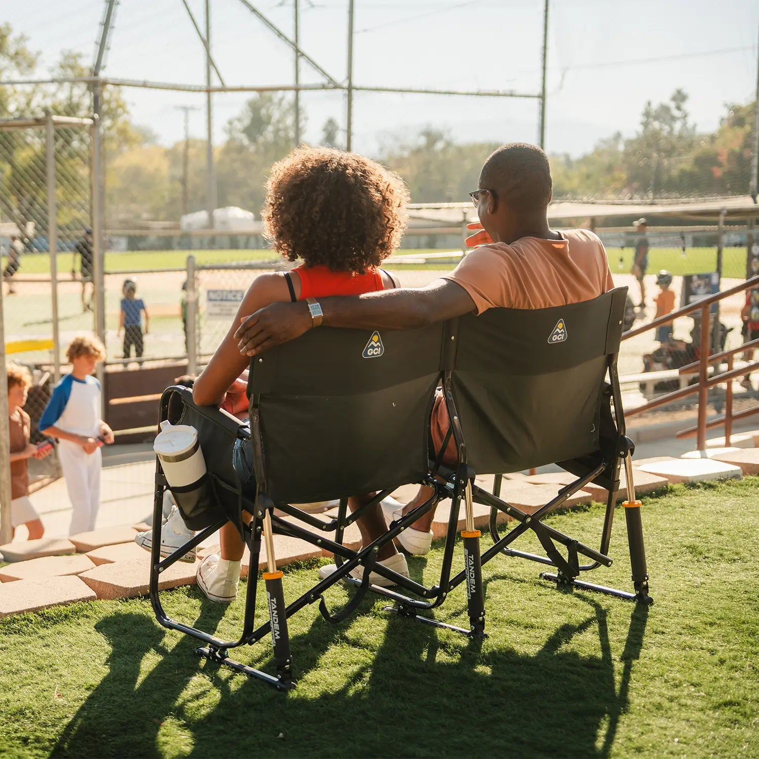 A couple sitting together on the Tandem Rocker while watching a baseball game.