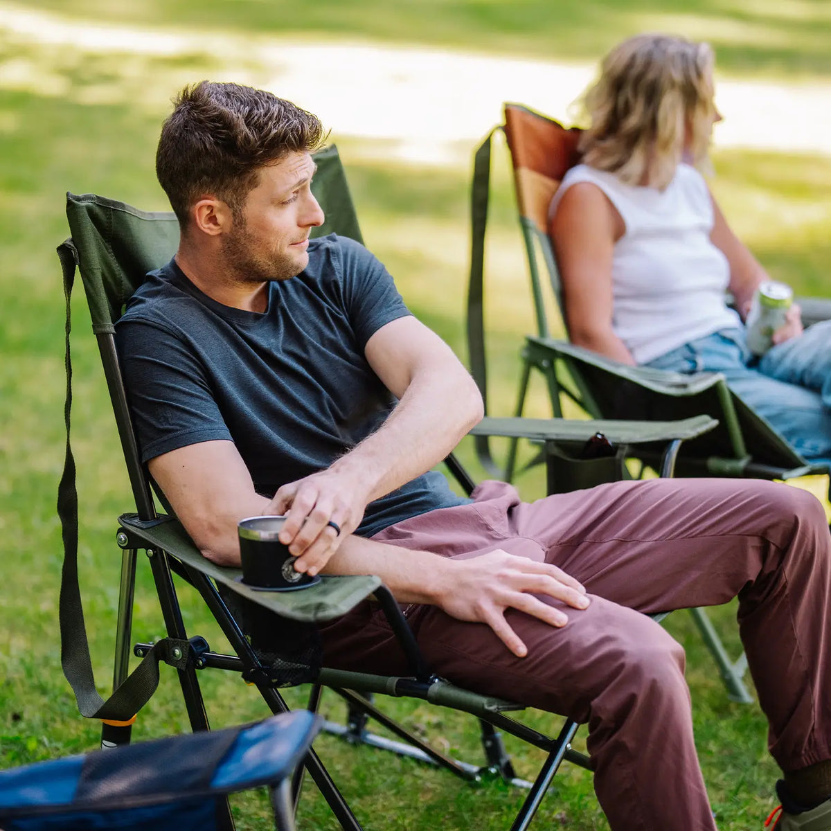A man sits in a Comfort Pro Rocker XL chair on a grassy lawn, holding a black mug. Another woman is seated in a folding chair in the background.