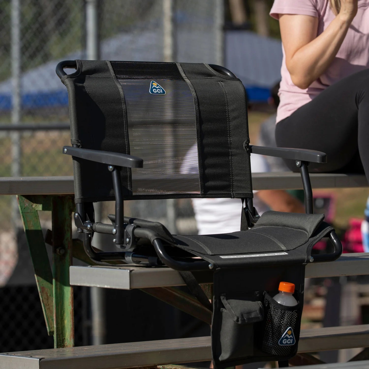 A dark charcoal Big Comfort Stadium Chair with Armrests 2.0 clips onto a bleacher bench, showing the mesh backrest and armrests.