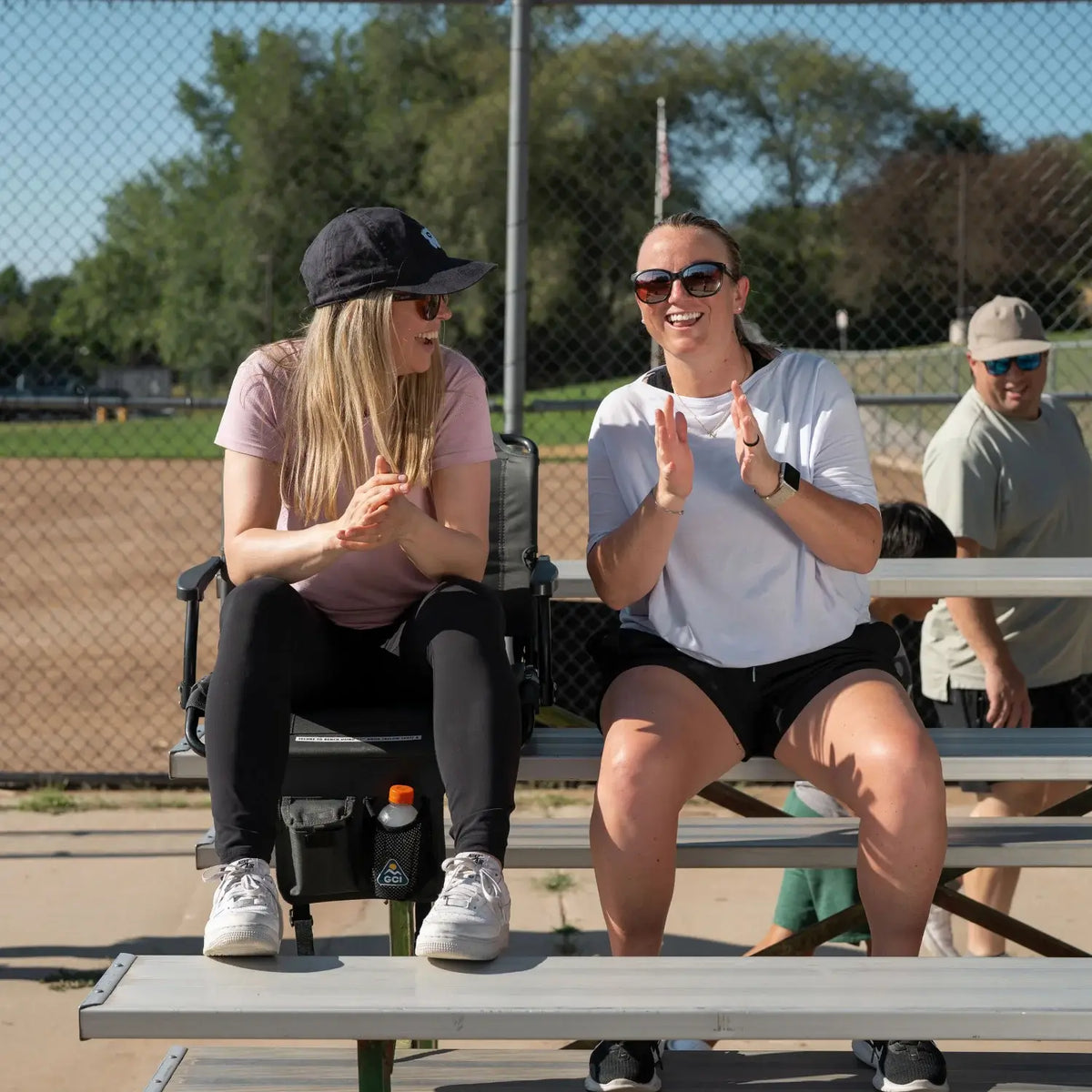 Two women laugh and clap while seated on bleachers, one in a dark charcoal Big Comfort Stadium Chair with Armrests 2.0.