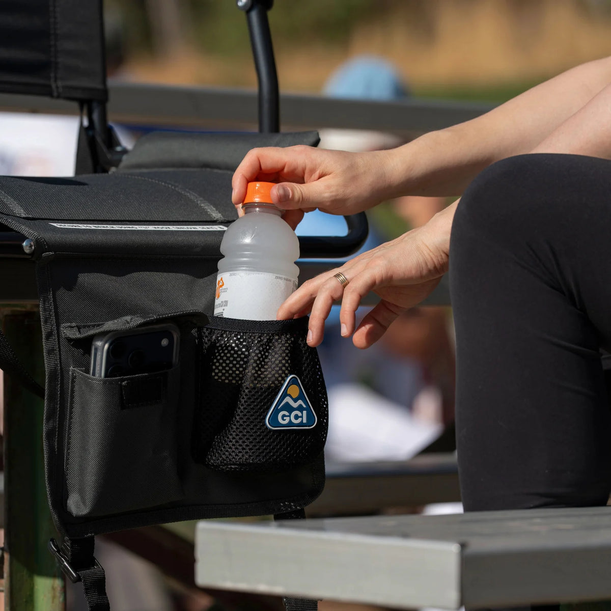 Close-up of hands placing a water bottle into the Pack & Port caddy on a dark charcoal Big Comfort Stadium Chair with Armrests 2.0, with a phone tucked in the side pocket.