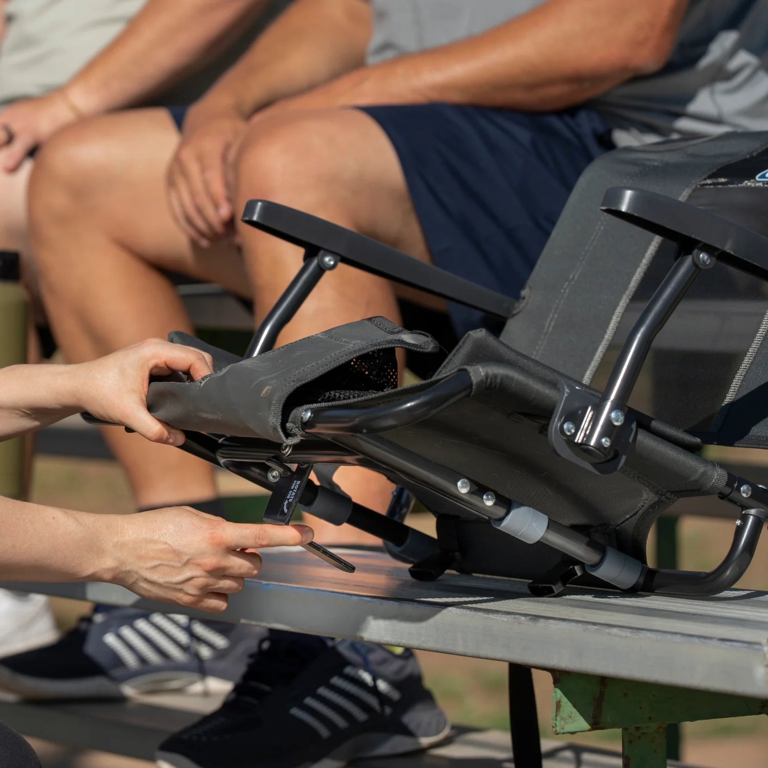 Close-up of hands adjusting the L-hook mechanism on a dark charcoal Big Comfort Stadium Chair with Armrests 2.0, secured beneath a bleacher bench.