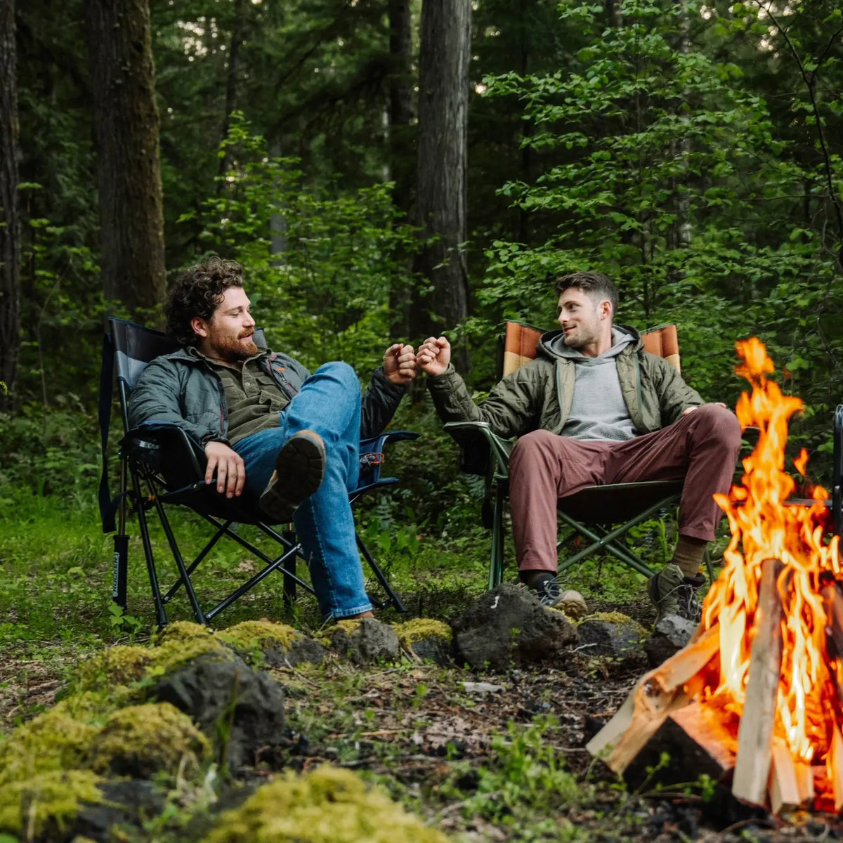 Two men sit in camp chairs, including an indigo Comfort Pro Insulated Rocker, fist-bumping beside a campfire in the woods.