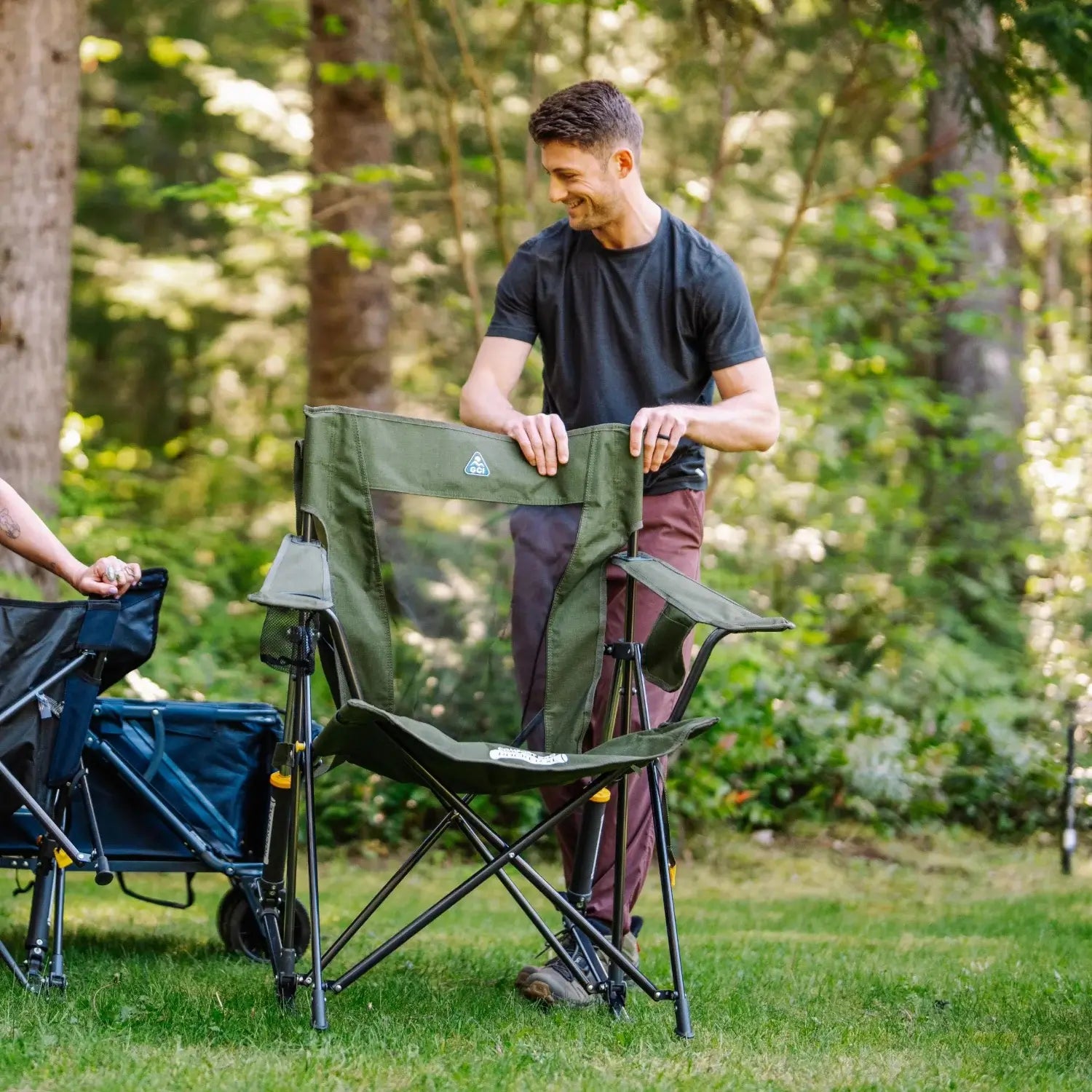 Man sets up a heathered olive Comfort Pro Rocker XL on grass near a wagon cart.
