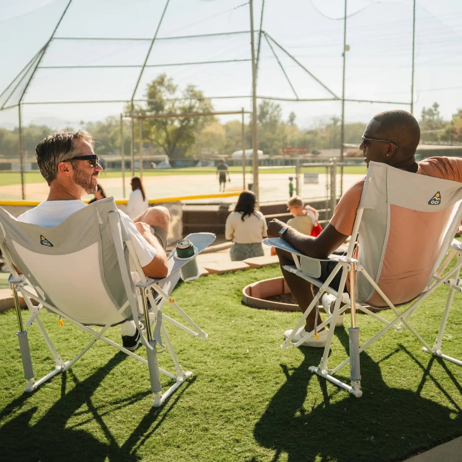 Two men chatting at a softball field in GCI Outdoor Comfort Pro Rocker XL chairs in Heathered Light Blue, rear angle view.