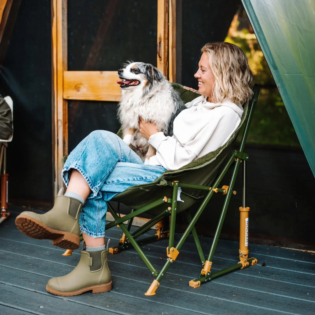 Woman relaxes in an olive Fleece Rocker with a dog on her lap, seated on a deck outside an A-frame cabin.