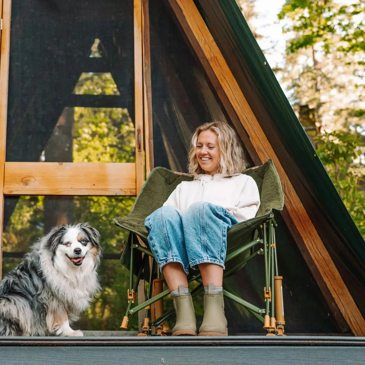 Woman sits in an olive Fleece Rocker with her feet up, smiling at a dog beside her on a wooden deck.