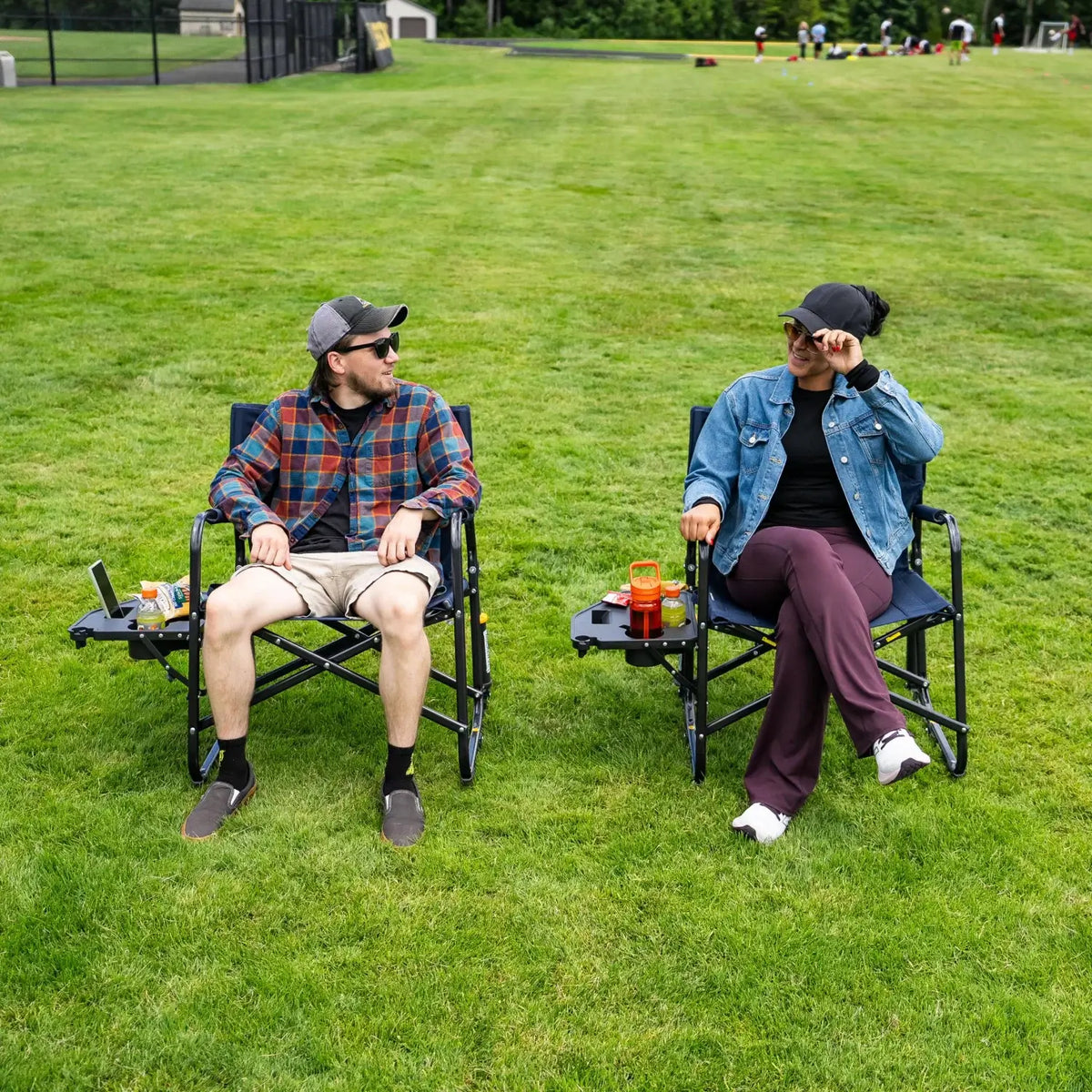 Two people sit facing each other in indigo Freestyle Rocker XL chairs with Side Table on a grass field.