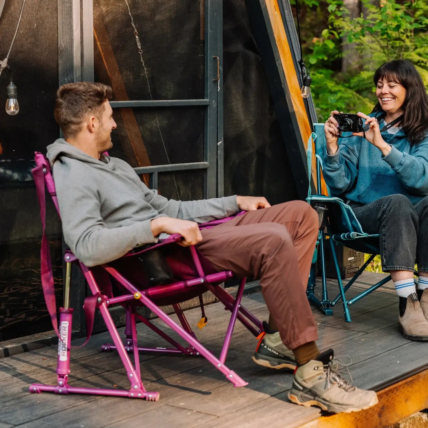 Man relaxes in a berry Kickback Insulated Rocker on a deck while a woman photographs him from a teal rocker.