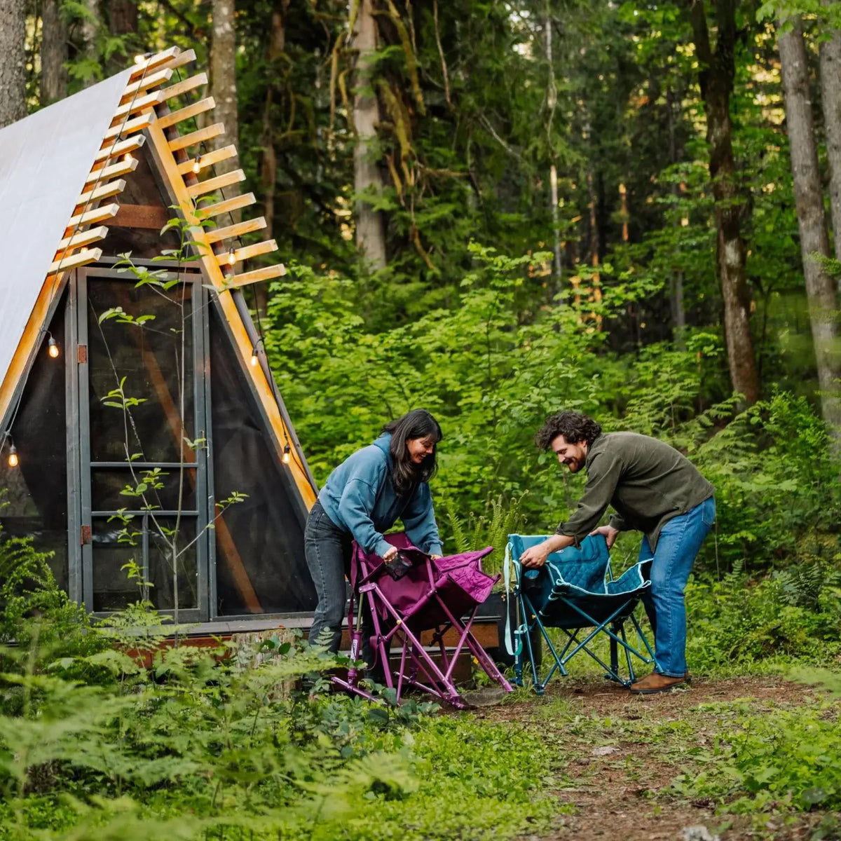 Two people fold up a berry and a teal Kickback Insulated Rocker outside an A-frame cabin surrounded by forest.