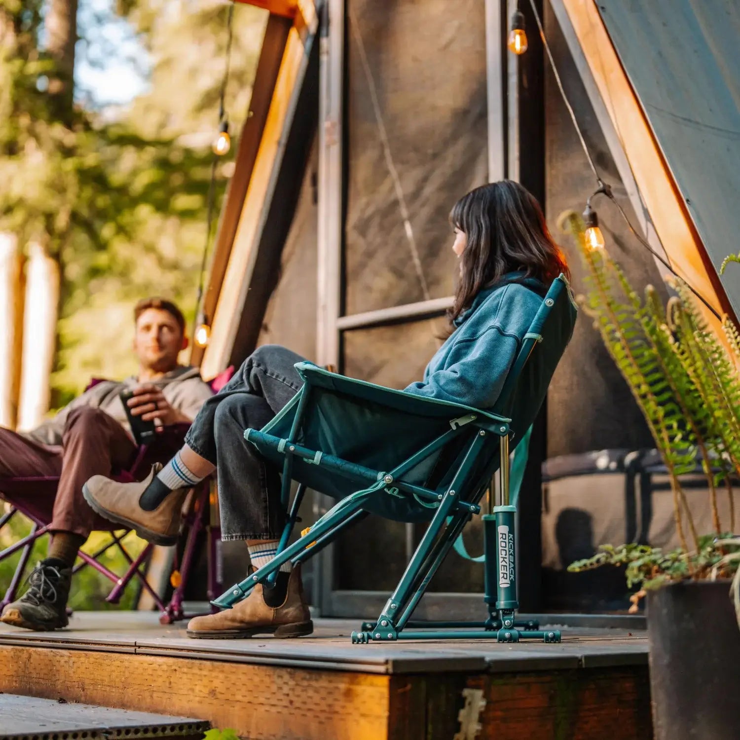 Woman sits in a teal Kickback Insulated Rocker on a wooden deck outside an A-frame cabin, with a man seated nearby.