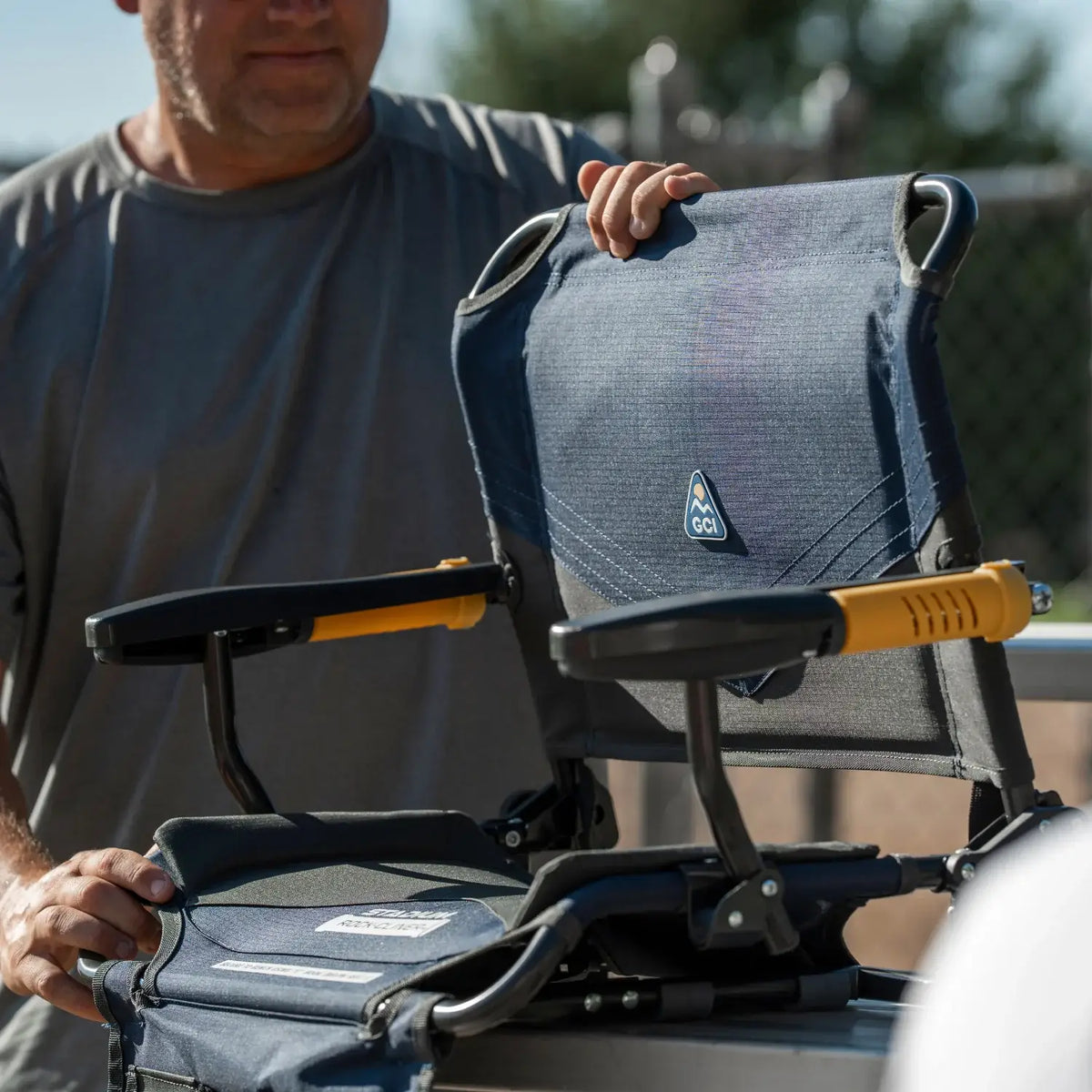 A man folds out the padded backrest of an indigo Stadium Rock-Cliner 2.0 bleacher seat, showing the armrests and bracket clamps.