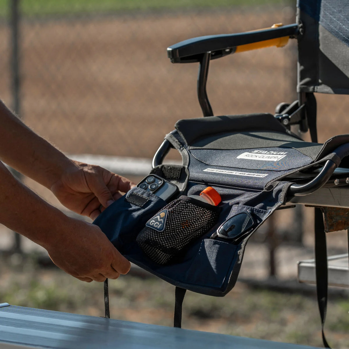 A hand opens the side pocket of an indigo Stadium Rock-Cliner 2.0, holding a phone, sunglasses, and a water bottle.