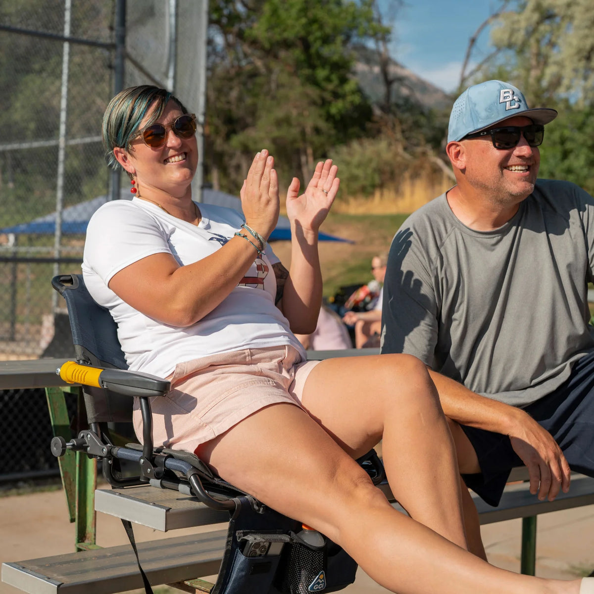 A woman claps while seated in an indigo Stadium Rock-Cliner 2.0 clamped to metal bleachers, with a man beside her also seated on the bleachers.