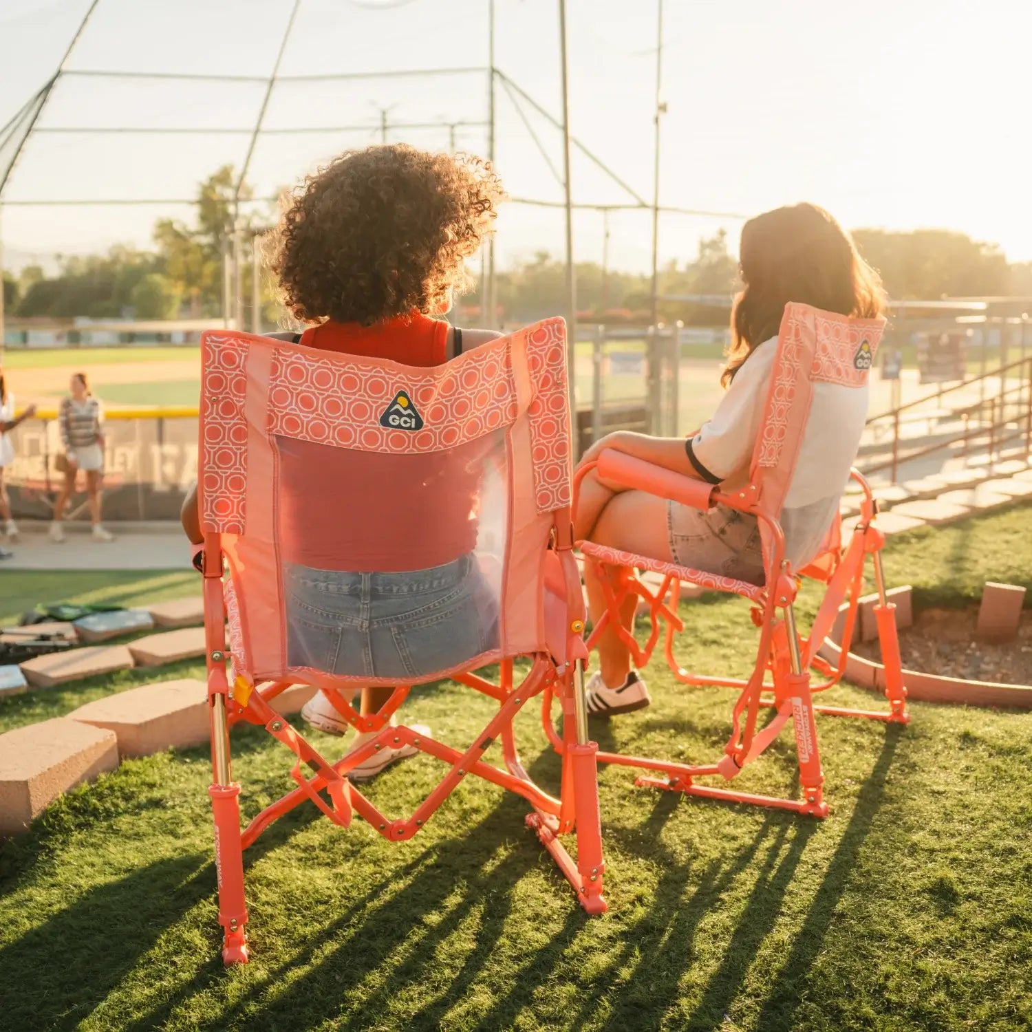 Two women watching a softball game from GCI Outdoor Stowaway Rockers in Coral Mosaic, rear view.
