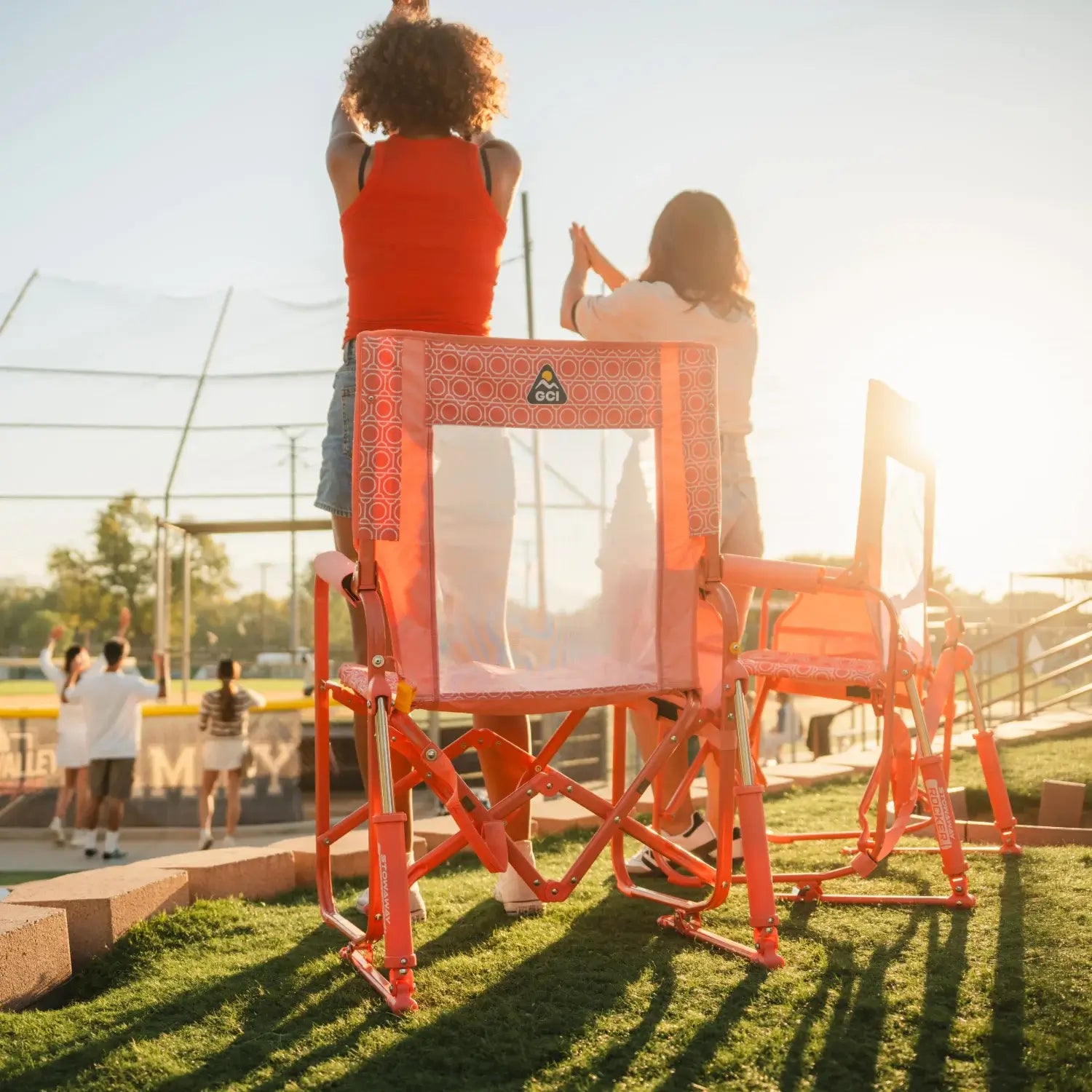 Two women cheering at a softball game beside GCI Outdoor Stowaway Rockers in Coral Mosaic, rear view.