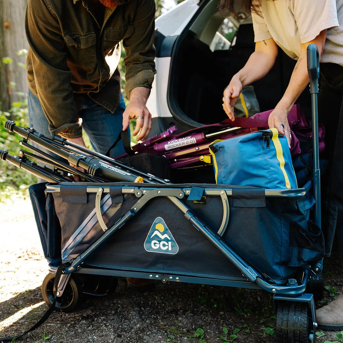 A GCI Go-Hauler being loaded with camping gear by two people outdoors. The wagon has a dark blue-gray fabric body, a silver metal frame, and black wheels, with a visible GCI logo on the front panel.