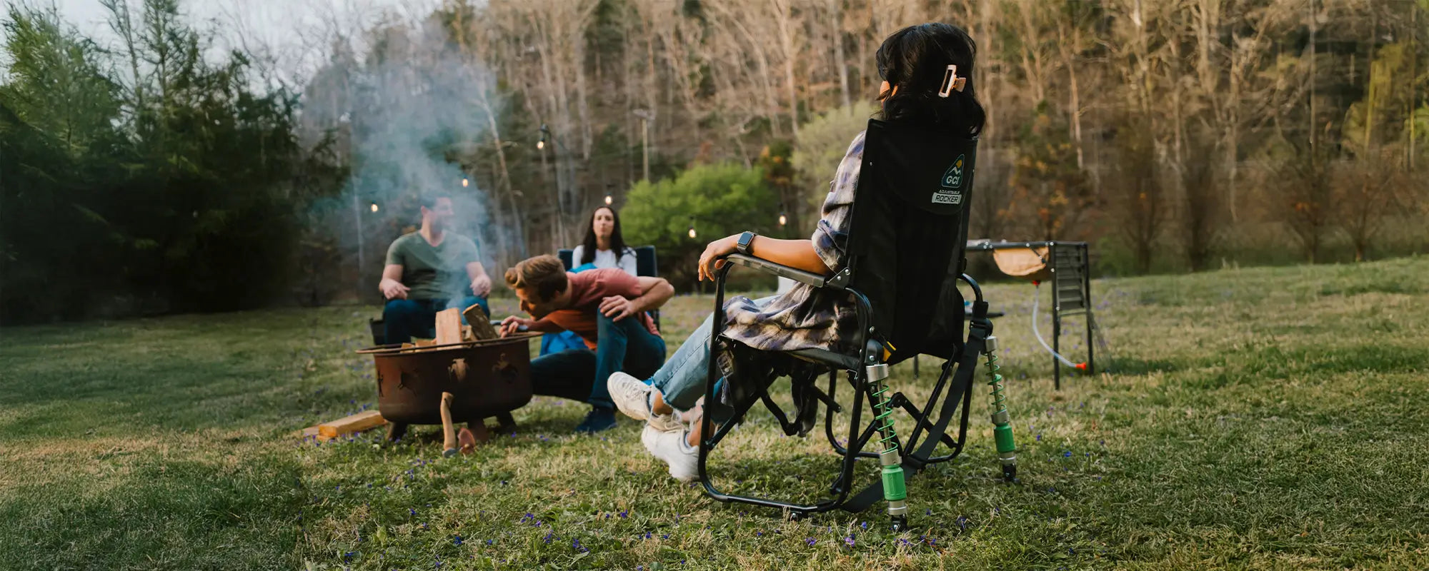A woman sitting next to a bonfire while in an adjustable rocker chair. 