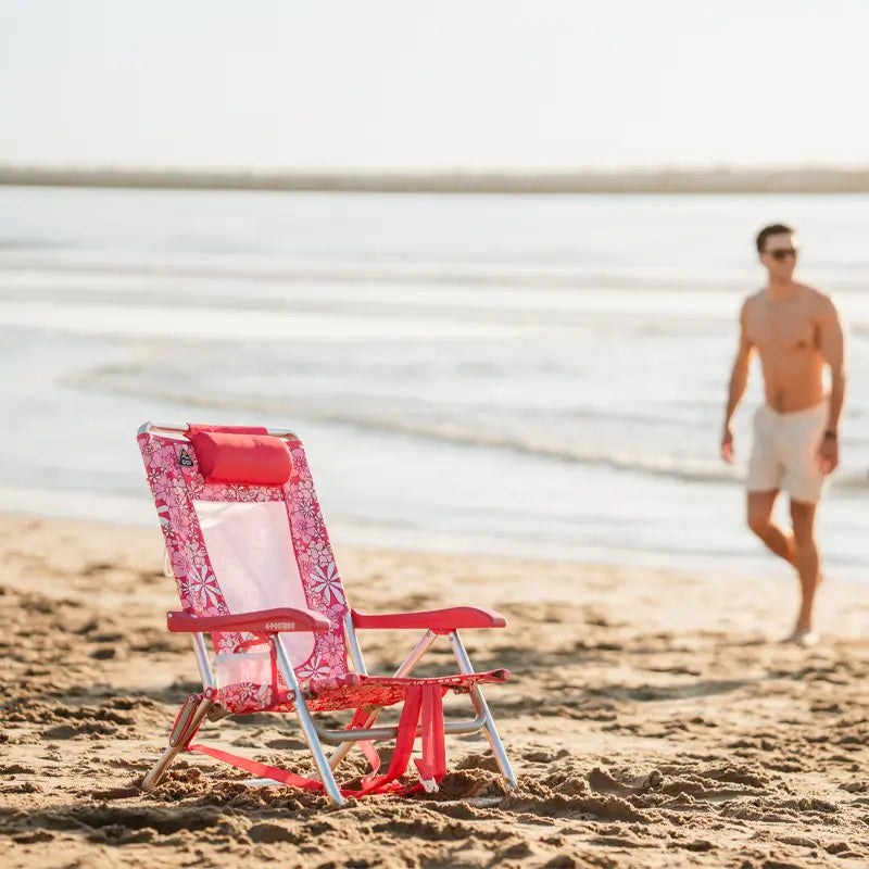 Pink Backpack Beach Chair with a floral pattern and pink headrest sitting on the sand with a man walking in the background.