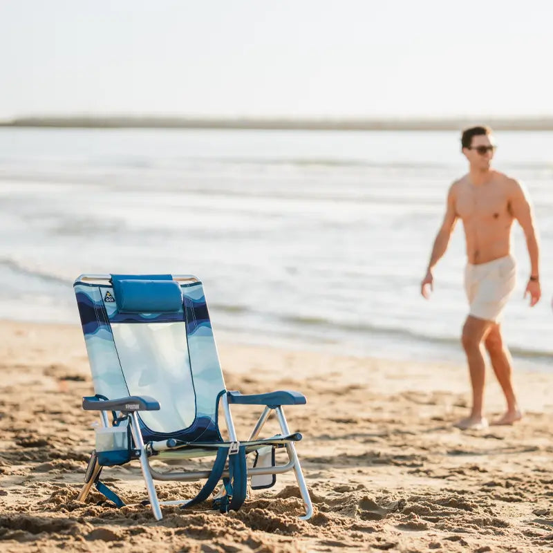 Blue wave-patterned Backpack Beach Chair sitting alone on the sand with the ocean and a man walking nearby.