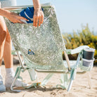 Woman placing a towel into the back pocket of a green floral Backpack Beach Chair with a cup in the holder.