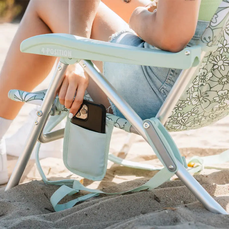 Woman sliding a phone into the side pouch of a green floral Backpack Beach Chair on the sand.