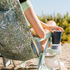 Woman sitting in a green floral Backpack Beach Chair holding a drink at the beach.