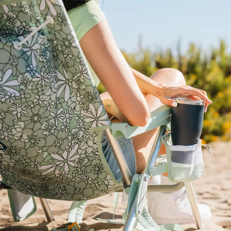 Woman sitting in a green floral Backpack Beach Chair holding a drink at the beach.