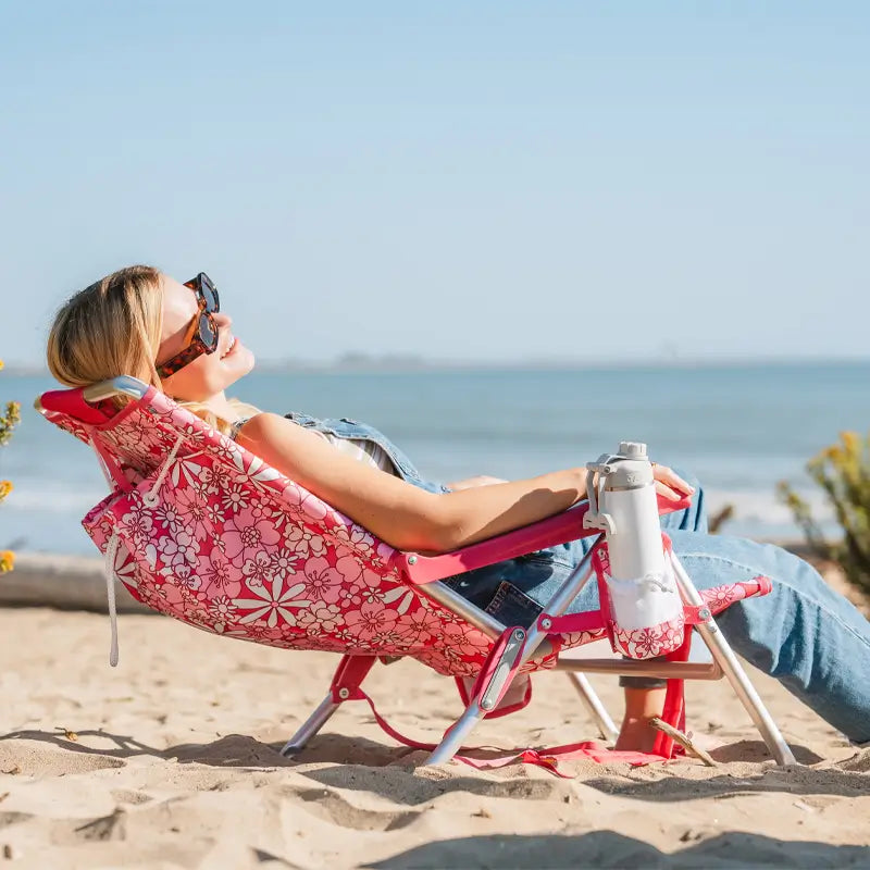 Woman reclining on a pink floral Backpack Beach Chair with a drink bottle secured in the cup holder.