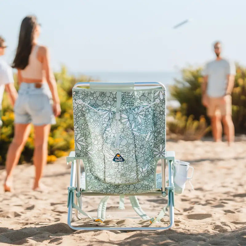 Green floral Backpack Beach Chair facing the ocean with people playing in the background.