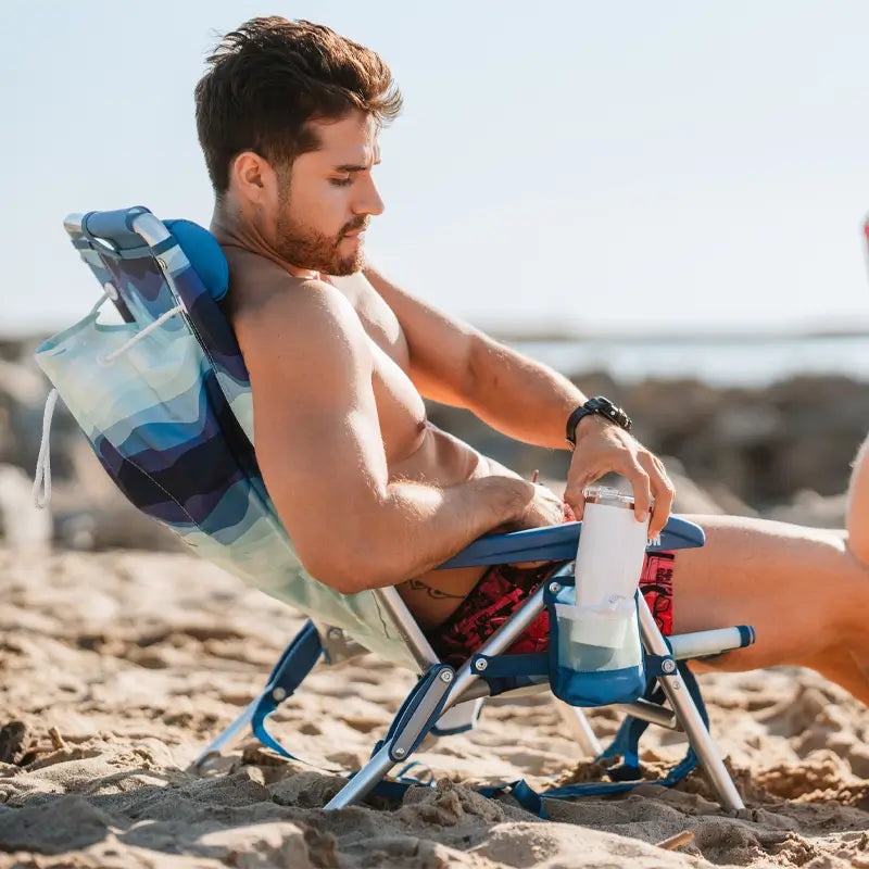 Shirtless man in red shorts sitting in a blue wave-patterned Backpack Beach Chair, holding a drink in the cup holder.