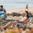 Woman reclining in a striped Backpack Beach Chair, smiling, with the ocean and rocks in the background.