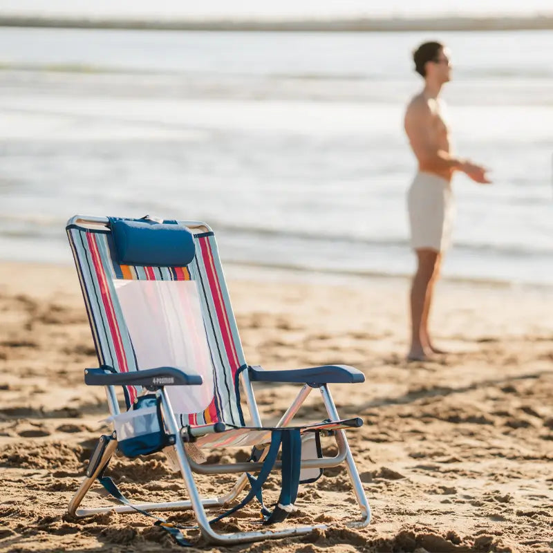 Striped Backpack Beach Chair with a blue headrest sitting alone on the sand with the ocean and a man walking in the background.