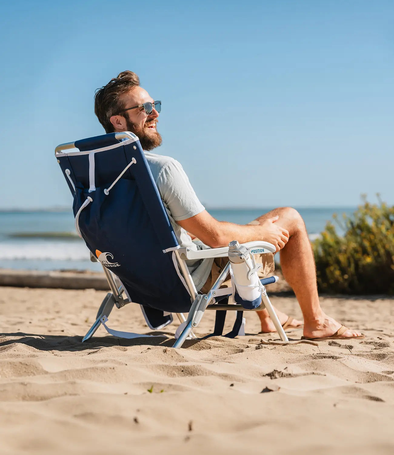 A man wearing sunglasses sits in a navy blue folding beach chair on a sandy beach with the ocean and blue sky in the background.