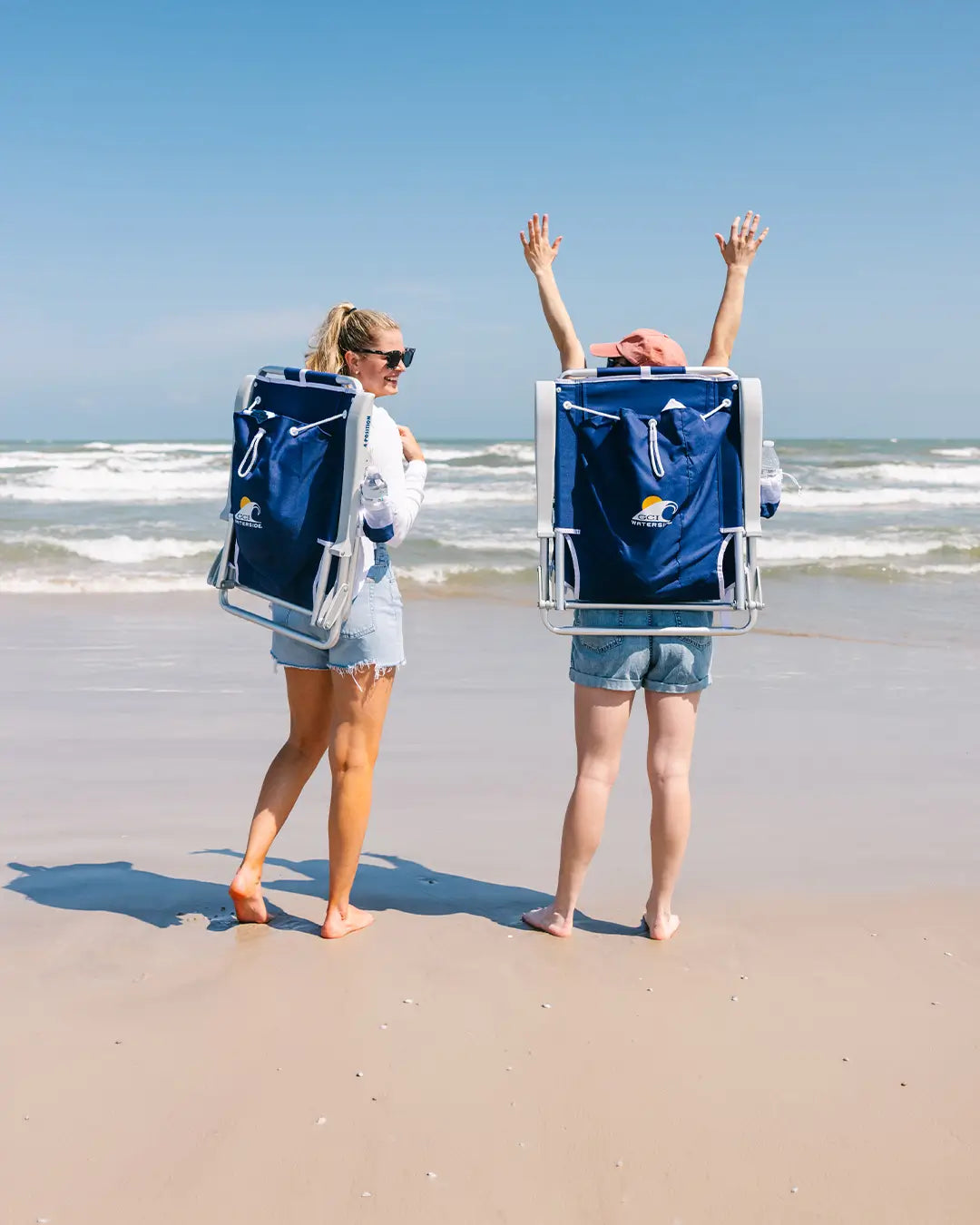 Two women walking on the beach with their nautical blue Backpack Beach chairs on their backs. 