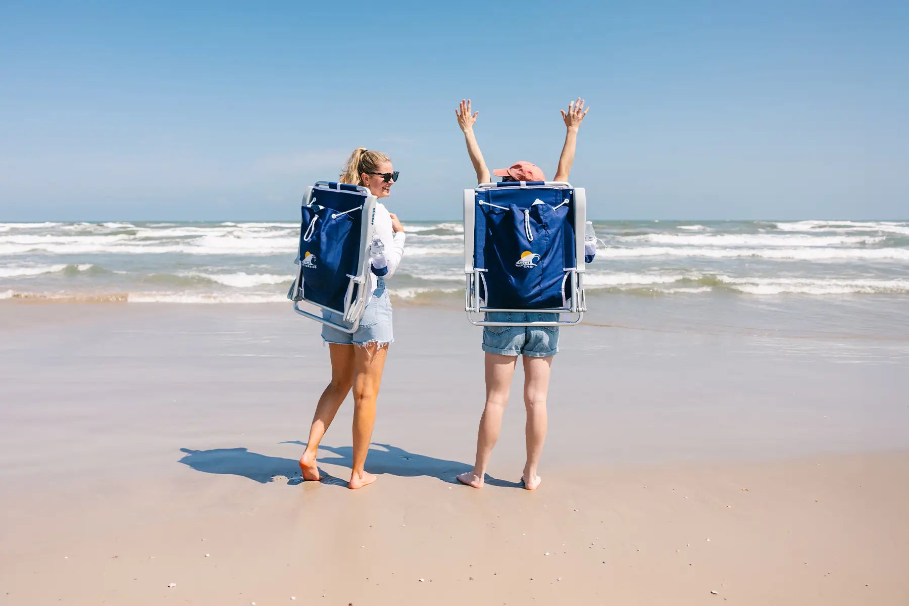 Two women cheering on a beach while carrying blue Backpack Beach Chairs. 
