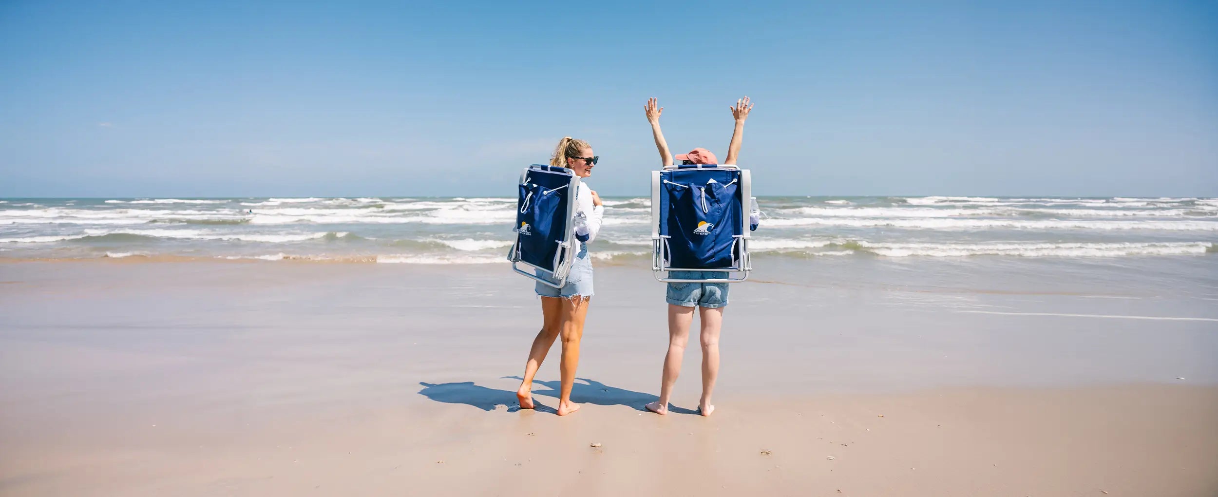 Two women cheering on a beach while carrying blue Backpack Beach Chairs. 