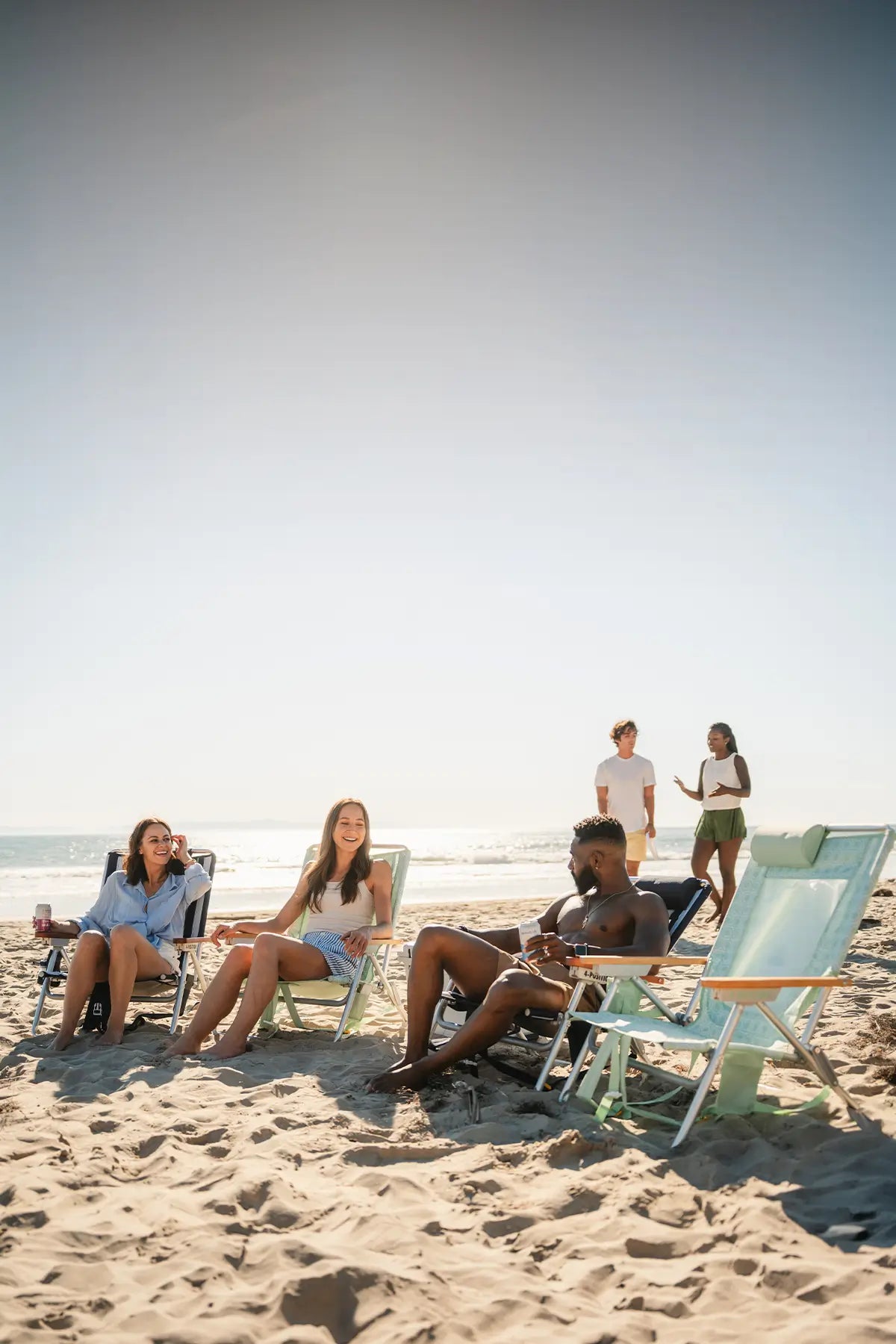 Several people relax in beach chairs on a sandy beach, with the ocean and a clear sky in the background.