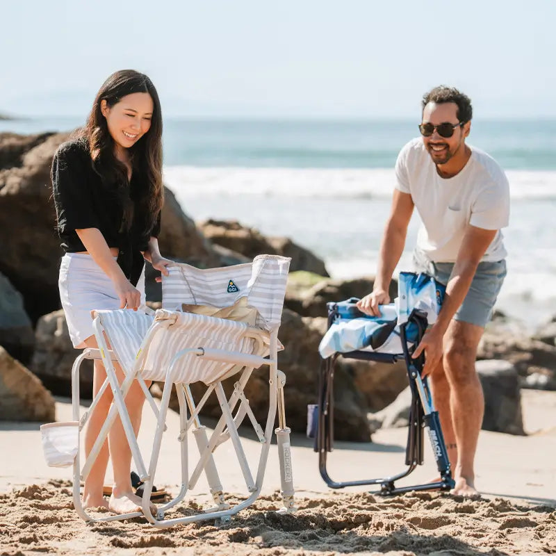 A woman in a black shirt and white shorts setting up a beige striped Beach Rocker chair while a man in sunglasses unfolds a blue chair nearby.