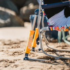 Close-up of a beach rocker on sandy ground with a person seated, showing the chair's metal frame and bright orange shocks in a beach setting.