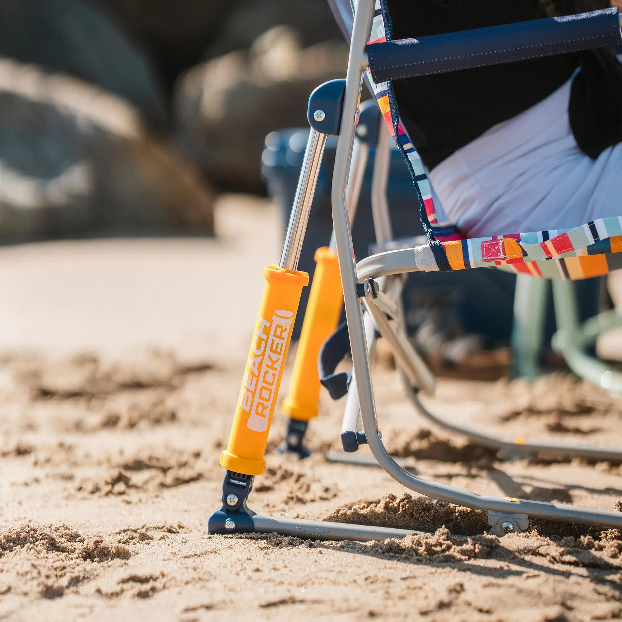 Close-up of a beach rocker on sandy ground with a person seated, showing the chair's metal frame and bright orange shocks in a beach setting.
